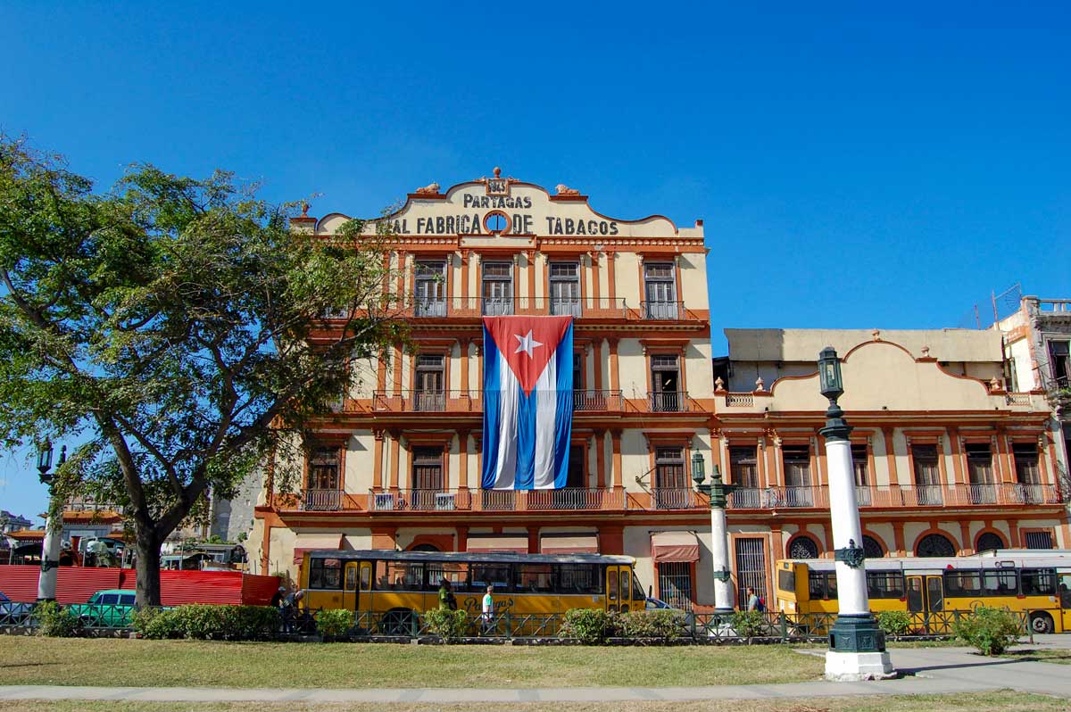 Real Fabrica de Tabacos Partagás, Havana – Photo: Shutterstock large old factory building with cuban flag behind city square